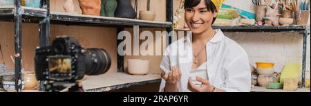 Joyeux jeune potier féminin asiatique parlant à l'appareil photo numérique flou tout en étant assis près des sculptures d'argile sur le rack dans l'atelier de céramique, argile sculptant pr Banque D'Images