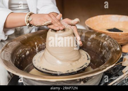 Vue recadrée de la jeune artiste féminine moulant de l'argile humide sur la roue de poterie tout en travaillant près du bol flou avec de l'eau et des outils dans l'atelier, studio de poterie Banque D'Images