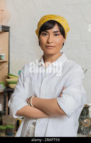 Portrait de jeune potier asiatique femelle en foulard et vêtements de travail croisant les bras et regardant la caméra tout en se tenant debout dans l'atelier de céramique, atelier de poterie Banque D'Images