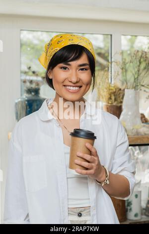 Portrait de jeune artisan asiatique souriant en foulard et vêtements de travail regardant la caméra et tenant le café pour aller tout en se tenant debout dans la poterie floue Banque D'Images