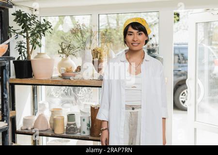 Portrait de jeune artisan asiatique souriant en vêtements de travail et foulard regardant la caméra tout en se tenant près de sculptures en céramique à l'arrière-plan dans l'art Banque D'Images