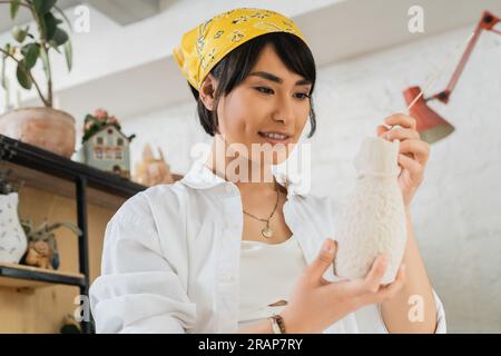 Souriant jeune artiste féminine asiatique en foulard et vêtements de travail tenant bâton en bois et sculpture en argile tout en travaillant dans la classe de poterie floue, poterie St Banque D'Images