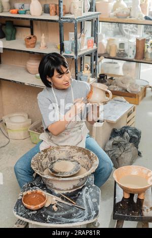 Vue à grand angle de la jeune femme asiatique artisan dans la peinture de tablier sur la cruche d'argile près de la roue de poterie, outils en bois et bol avec de l'eau dans l'atelier de céramique, Banque D'Images