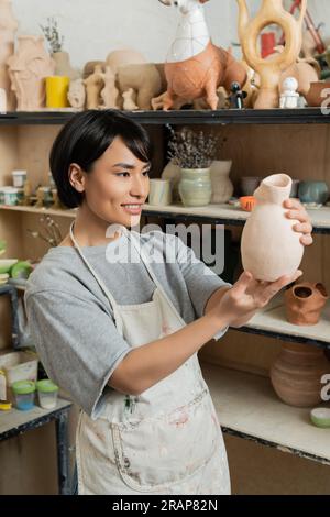 Jeune artisan asiatique souriant en tablier et vêtements de travail regardant la sculpture en céramique tout en se tenant près du rack dans l'atelier de céramique floue à Backgrou Banque D'Images