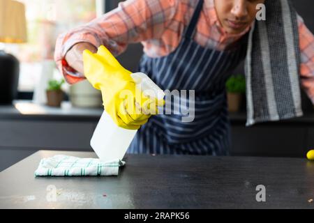 Section médiane de l'homme biracial portant un tablier et des gants en caoutchouc nettoyant le comptoir dans la cuisine ensoleillée. Nettoyage, hygiène, mode de vie sain et lif domestique Banque D'Images