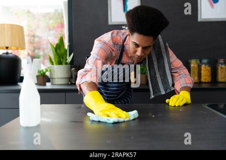Homme biracial concentré portant un tablier et des gants en caoutchouc nettoyant le comptoir dans la cuisine ensoleillée. Nettoyage, hygiène, mode de vie sain et vie domestique, una Banque D'Images