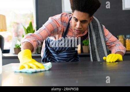 Homme biracial concentré portant un tablier et des gants en caoutchouc nettoyant le comptoir dans la cuisine ensoleillée. Nettoyage, hygiène, mode de vie sain et vie domestique, una Banque D'Images
