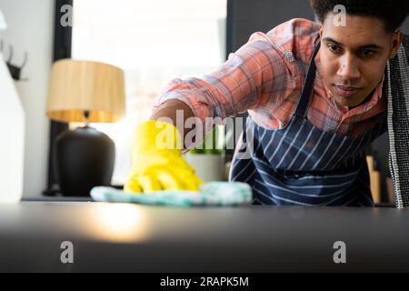 Homme biracial concentré portant un tablier et des gants en caoutchouc nettoyant le comptoir dans la cuisine ensoleillée. Nettoyage, hygiène, mode de vie sain et vie domestique, una Banque D'Images