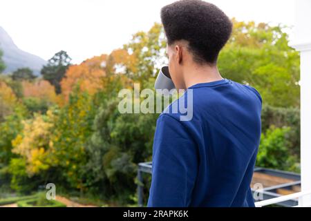 Vue arrière de l'homme biracial réfléchi debout sur le balcon buvant du café et profitant de la vue sur le jardin. Temps libre, mode de vie et vie domestique, unalter Banque D'Images