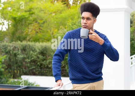 Homme biracial réfléchi debout sur le balcon buvant du café et regardant loin. Temps libre, mode de vie et vie domestique, inchangés. Banque D'Images