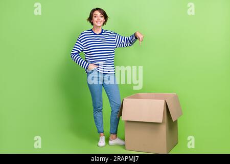 Photo pleine longueur de la femme surjoyée porter un pantalon en denim chemise marin indiquant à la main de boîte dans la poche isolé sur fond de couleur verte Banque D'Images