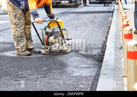 Un travailleur des services routiers pilotant de l'asphalte frais sur un tronçon de route avec une vieille plaque vibrante d'essence usée autour d'un regard d'égout. Espace de copie. Banque D'Images