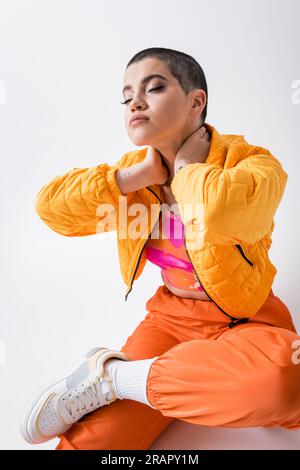 vêtements d'extérieur, avant-garde de la mode, jeune femme tatouée dans des vêtements colorés assis avec les yeux fermés sur fond gris, style urbain, individualisme, vibrant Banque D'Images