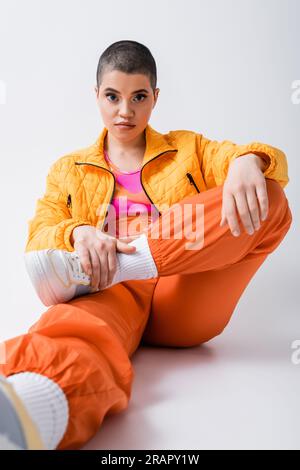 vêtements d'extérieur, déclaration de mode, jeune femme tatouée dans des vêtements colorés assis et regardant la caméra sur fond gris, style urbain, individualisme, v Banque D'Images