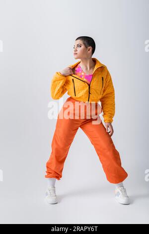 vêtements d'extérieur, tenue décontractée, tatouée et jeune femme avec les cheveux courts tirant veste gonflante jaune sur fond gris, mode urbaine, jeunesse vibrante, tren Banque D'Images