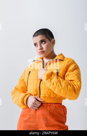 vêtements d'extérieur, tatoués et jeune femme avec cheveux courts zippant veste jaune bouffante sur fond gris, mode urbaine, jeunesse vibrante, tenue tendance, styl Banque D'Images