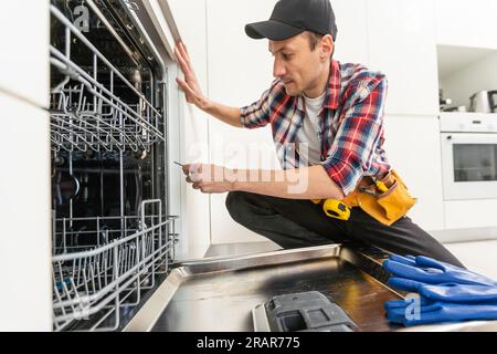 Tous les instruments sont inclus. Technicien mâle debout près de lave-vaisselle à l'aide d'un tournevis dans la cuisine avec des instruments Banque D'Images