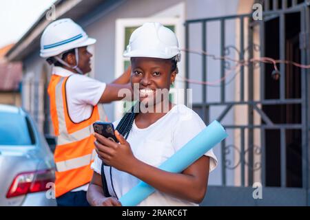 portrait d'une heureuse africaine nigériane heureuse africaine nigériane ingénieure portant un casque blanc, tenant des plans et utilisant smartphone, fil Banque D'Images