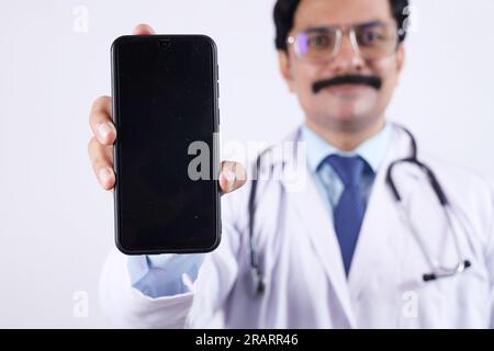 Beau portrait de médecin indien heureux en uniforme portant stéthoscope et spécifications montrant l'écran du téléphone en main. Affichage de l'application mobile. Banque D'Images