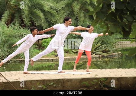 Jeune homme en forme s'exerçant et faisant des poses de yoga dans un environnement vert tôt le matin près du plan d'eau avec des enfants. Mode de vie sain. Journée internationale du yoga. Banque D'Images