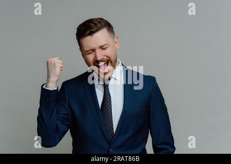 Homme d'affaires prospère célèbre, serre le poing, s'exclame dans la joie, habillé formellement, satisfait après l'accord. Fond gris. Félicitations, je l'ai fait ! Banque D'Images