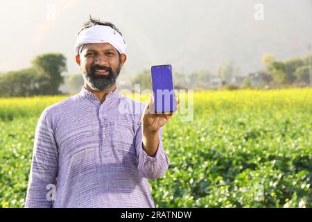 Portrait d'un fermier heureux rural indien debout dans le champ de moutarde portant kurta pyjama en été. Agriculteur montrant l'application mobile sur l'écran du smartphone. Banque D'Images
