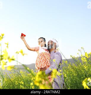 Agriculteurs ruraux indiens heureux. Père et fille dans le champ de moutarde jouant avec des avions en papier. Fille rêve de voler à l'étranger. Hôtesse de l'air. Banque D'Images
