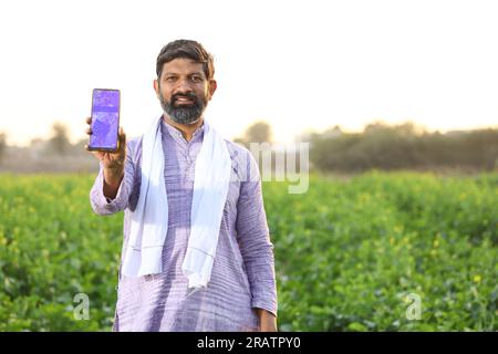 Portrait d'un fermier heureux rural indien debout dans le champ de moutarde portant kurta pyjama en été. Agriculteur montrant l'application mobile sur l'écran du smartphone. Banque D'Images