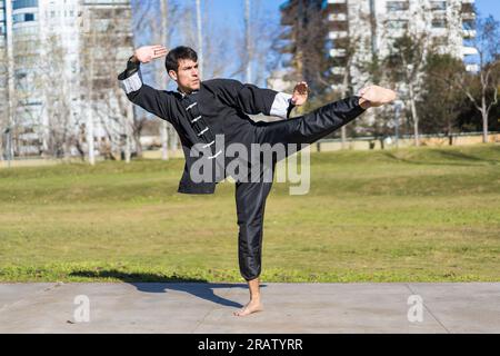 Jeune combattant athlétique d'arts martiaux pratiquant des coups de pied dans un parc public portant l'uniforme de kung fu. Banque D'Images