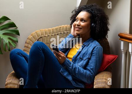 Heureuse femme biracial avec casque écoutant de la musique en utilisant smartphone à la maison. Relaxation, style de vie, communication et vie domestique, inchangés. Banque D'Images