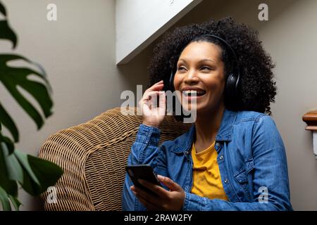 Heureuse femme biracial avec casque écoutant de la musique en utilisant smartphone à la maison. Relaxation, style de vie, communication et vie domestique, inchangés. Banque D'Images