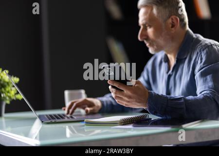 Homme d'affaires mûr souriant tenant smartphone assis dans le bureau. directeur général d'âge moyen utilisant des applications mobiles de téléphone portable et un ordinateur portable. Technologie numérique a Banque D'Images