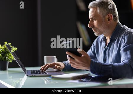 Homme d'affaires mûr souriant tenant smartphone assis dans le bureau. directeur général d'âge moyen utilisant des applications mobiles de téléphone portable et un ordinateur portable. Technologie numérique a Banque D'Images