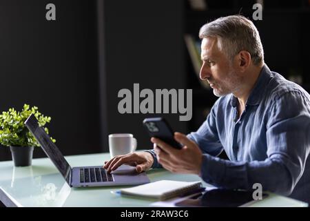 Homme d'affaires mûr souriant tenant smartphone assis dans le bureau. directeur général d'âge moyen utilisant des applications mobiles de téléphone portable et un ordinateur portable. Technologie numérique a Banque D'Images