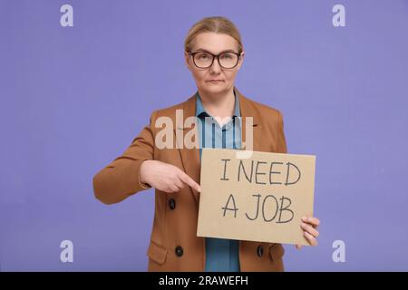 Femme âgée sans emploi pointant du doigt le signe en carton avec la phrase I Need A Job sur fond violet Banque D'Images