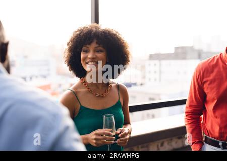 Joyeuse femme d'affaires décontractée biracial buvant du champagne et parlant avec des collègues au bureau. Affaires occasionnelles, travail d'équipe, liens, succès et célébration Banque D'Images