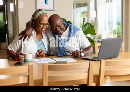 Heureux senior couple afro-américain embrassant, utilisant un ordinateur portable et payant des factures à la maison. Style de vie senior, finance, vie domestique et retraite, unalte Banque D'Images