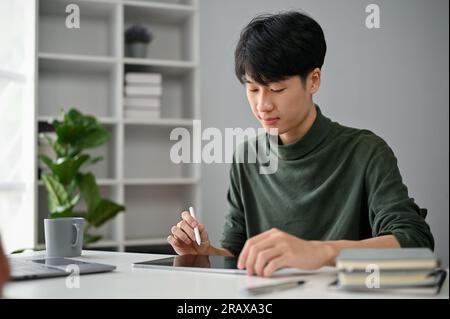 Un jeune homme asiatique concentré et intelligent dans des vêtements décontractés utilisant sa tablette numérique pour gérer ses tâches à son bureau dans le bureau. Banque D'Images