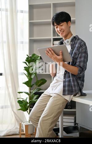 Un portrait d'un jeune homme asiatique heureux et beau dans une chemise en flanelle utilisant sa tablette numérique dans son bureau moderne. Banque D'Images