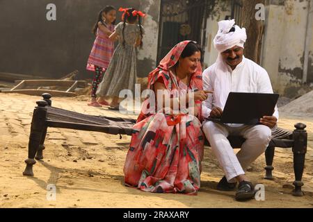 Heureuse famille indienne rurale dans le village. Couple rural assis ensemble souriant sur lit bébé devant leur maison dans la cour avant en utilisant un ordinateur portable pour les paiements en ligne. Banque D'Images