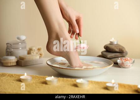 Femme trempant son pied dans un bol avec de l'eau et des pétales sur plancher en bois blanc, gros plan. Procédé de pédicure Banque D'Images