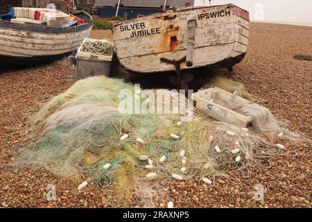 Deux petits vieux bateaux de pêche en bois sur la plage de galets à Aldeburgh, Suffolk. Royaume-Uni avec un tas de filets de chalutiers fins et des flotteurs couchés sur les pierres Banque D'Images