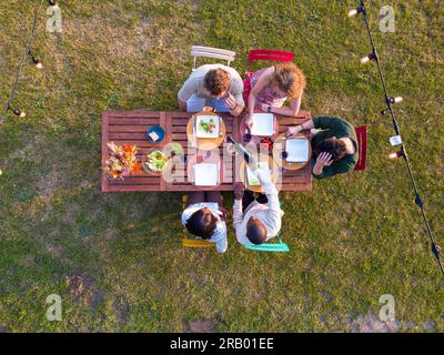 Une prise de vue aérienne captivante capturant un groupe de jeunes amis de diverses ethnies réunis autour d'une table, dégustant une variété de plats et de boissons lors d'une fête d'été. L'image englobe magnifiquement les loisirs, la célébration et le concept de convivialité pendant les vacances et les pique-niques. Vue aérienne de Summer Garden Party : amis multiraciaux profitant de la nourriture à l'extérieur. Photo de haute qualité Banque D'Images