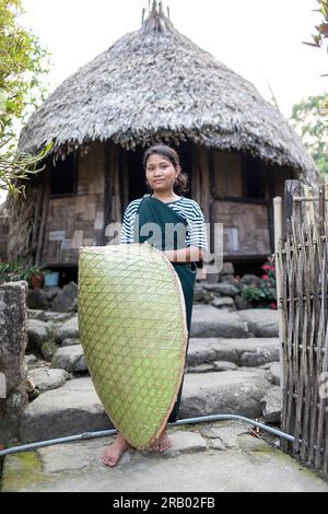 Fille de la tribu khasi en vêtements traditionnels portant une couverture de pluie traditionnelle en bambou, devant une maison khasi traditionnelle, Meghalaya, Inde Banque D'Images