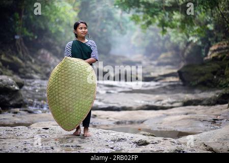 Fille de la tribu khasi dans des vêtements traditionnels portant une couverture de pluie traditionnelle en bambou, debout dans le lit rocheux de la rivière, Meghalaya, Inde Banque D'Images