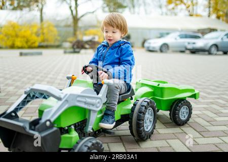 Adorable garçon en bas âge chevauchant son tracteur jouet dans une ville sur la soirée ensoleillée d'automne. Jeune enfant roulant sur un rouleau. Loisirs actifs et sports de plein air pour les enfants Banque D'Images