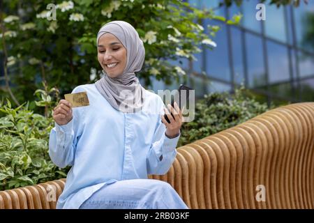 Jeune belle femme dans le hijab avec le téléphone dans les mains et la carte de crédit bancaire, musulman extérieur immeuble de bureaux réservant en ligne et commandant des vêtements, femme arabe sur les achats en ligne souriant, client satisfait. Banque D'Images