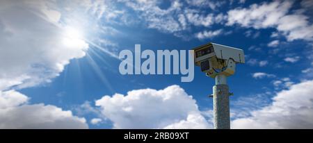 Gros plan d'une caméra de vitesse moderne (caméra de surveillance de la vitesse du trafic) contre un ciel bleu clair avec des nuages et des rayons de soleil. Banque D'Images