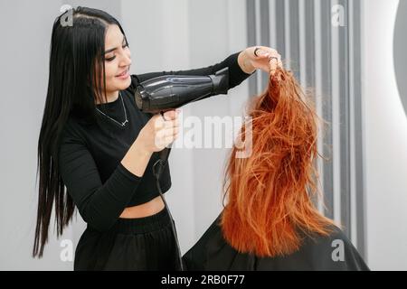Femme coiffeur fait coiffer avec sèche-cheveux au client dans le salon de beauté en face du miroir. Une femme aux cheveux rouges est assise dans un salon de coiffure. Styliste professionnelle fait la coiffure. Banque D'Images