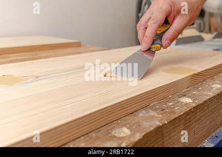 Couvrir les nœuds sur une planche de bois avec du mastic. Spatule, mastic, planche de bois. Les mains de Carpenter. Banque D'Images